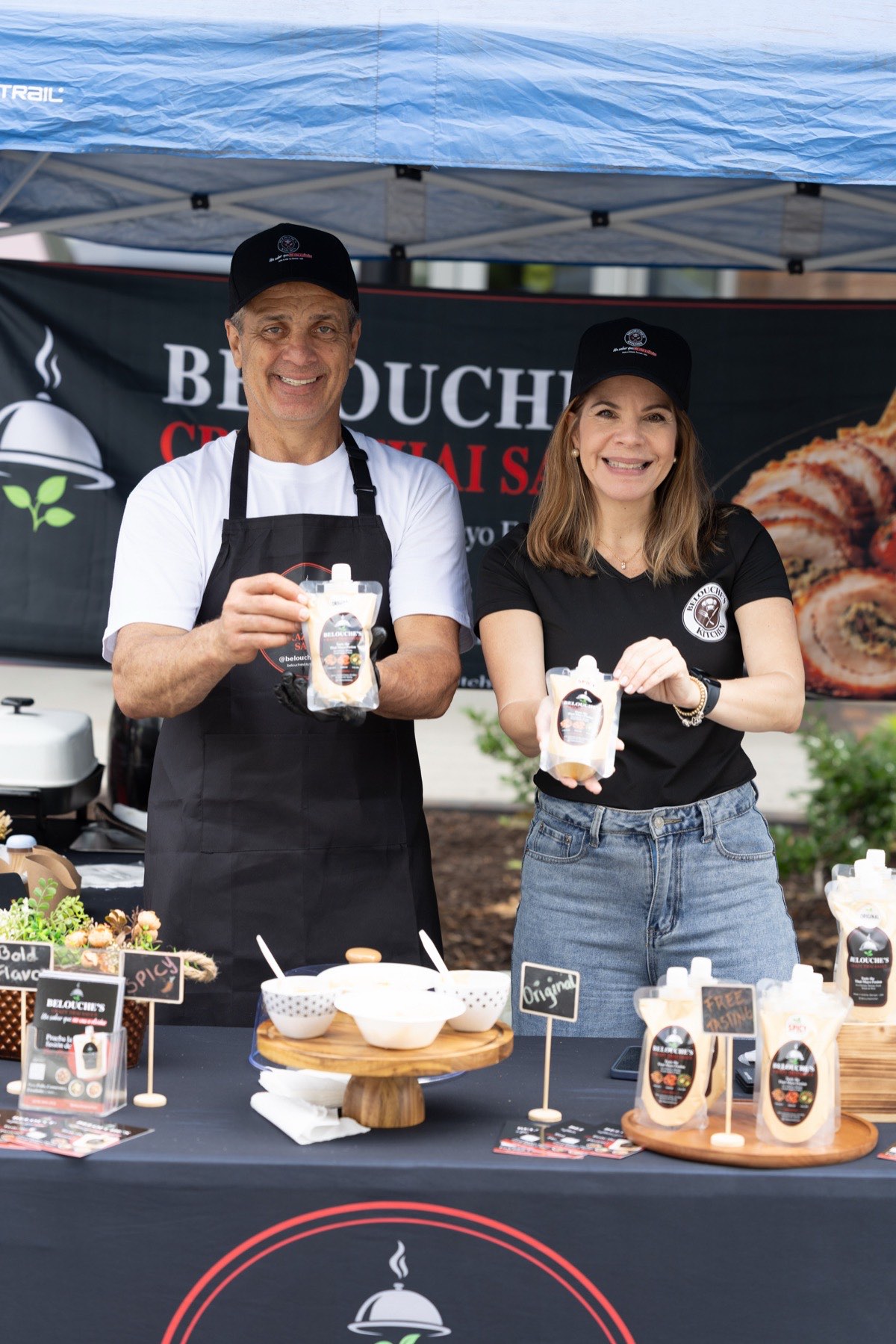 Belouche's Kitchen at their farmers market stand: two people in branded caps and aprons each holding a pouch of Crazy Thai Sauce, with Original and Spicy set out for tasting.