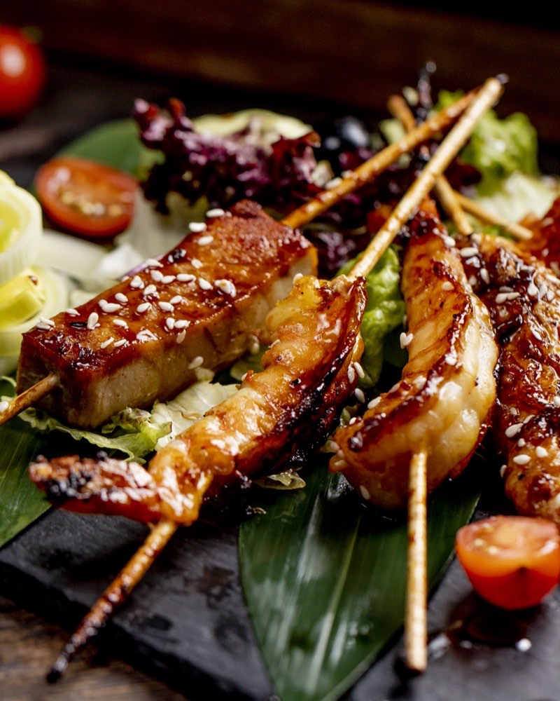 Glazed chicken skewers with sesame seeds on a slate board with salad leaves and tomato.